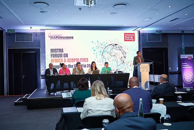 A conference panel sits on a stage in a hotel meeting room, with speakers seated at a table and one speaker standing at a podium. The screen behind them displays the MISTRA Forum on Africa & Geopolitics and Friedrich-Ebert-Stiftung logos. Attendees sit at tables facing the stage.