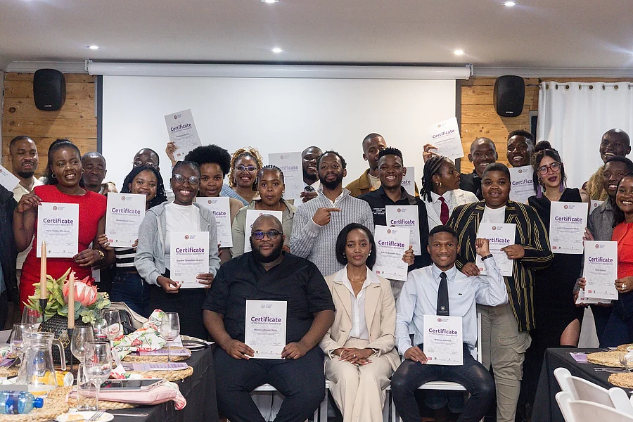 A group of  25 Fort Hare Activist School 2025 cohort participants pose together indoors, smiling and holding certificates of participation after completing a programme. Several participants raise their certificates toward the camera, while others sit in the front row.