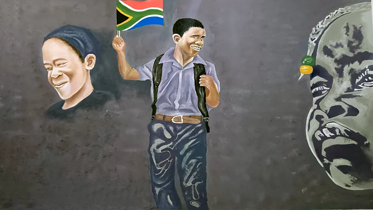Mural showing a joyful boy in a grey shirt and dark trousers, wearing a backpack with visible straps and a belt, smiling broadly while holding up a small South African flag. To his left is a softly rendered portrait of a smiling woman wearing a headscarf; to his right, a partial grayscale portrait of an older man's face appears mid-expression. The overall palette is muted grey apart from the boy's flag and clothing.