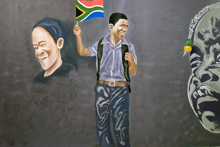 Mural showing a joyful boy in a grey shirt and dark trousers, wearing a backpack with visible straps and a belt, smiling broadly while holding up a small South African flag. To his left is a softly rendered portrait of a smiling woman wearing a headscarf; to his right, a partial grayscale portrait of an older man's face appears mid-expression. The overall palette is muted grey apart from the boy's flag and clothing.