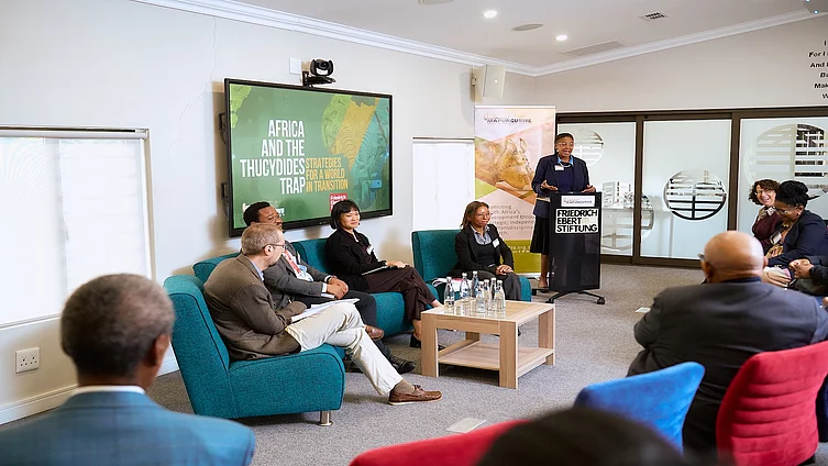 A speaker stands at a podium addressing an audience during a public policy panel discussion. Four panelists are seated on teal armchairs beside a coffee table with water bottles, while attendees listen from the audience. A large presentation screen displays the title, "Africa and the Thucydides Trap: Strategies for a World in Transition," and banners for the event organizers stand behind the podium, indicating a forum on international relations and Africa's role in a changing global order.