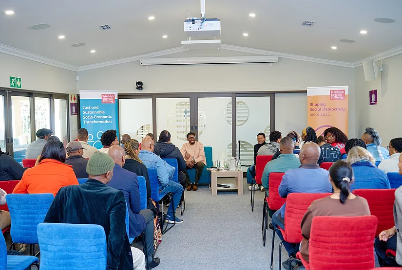 Audience seated in rows of red and blue chairs at an indoor event, facing two men seated in armchairs at the front of the room in a conversational setup, with a low wooden table between them. Two Friedrich Ebert Stiftung banner stands flank the seating area, reading 'Just and Sustainable Socio-Economic Transformation' and 'Shaping Social Democracy. Since 1925.