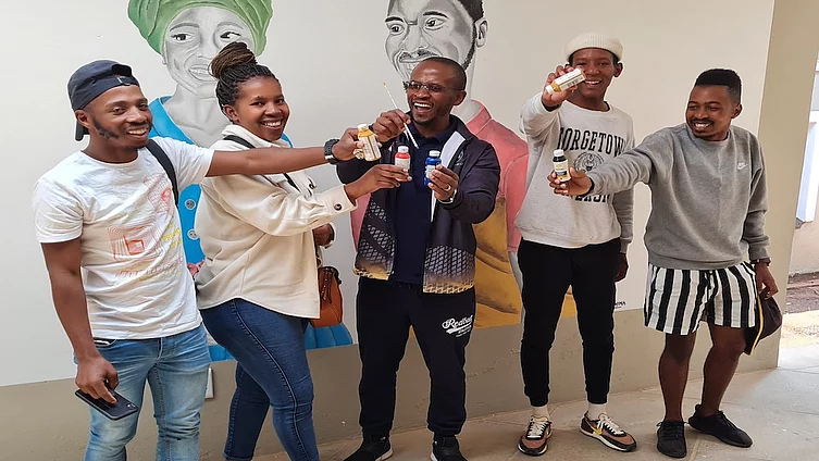 Group of five young people standing indoors in front of a mural featuring handpainted portraits, smiling and holding up small paint bottles.