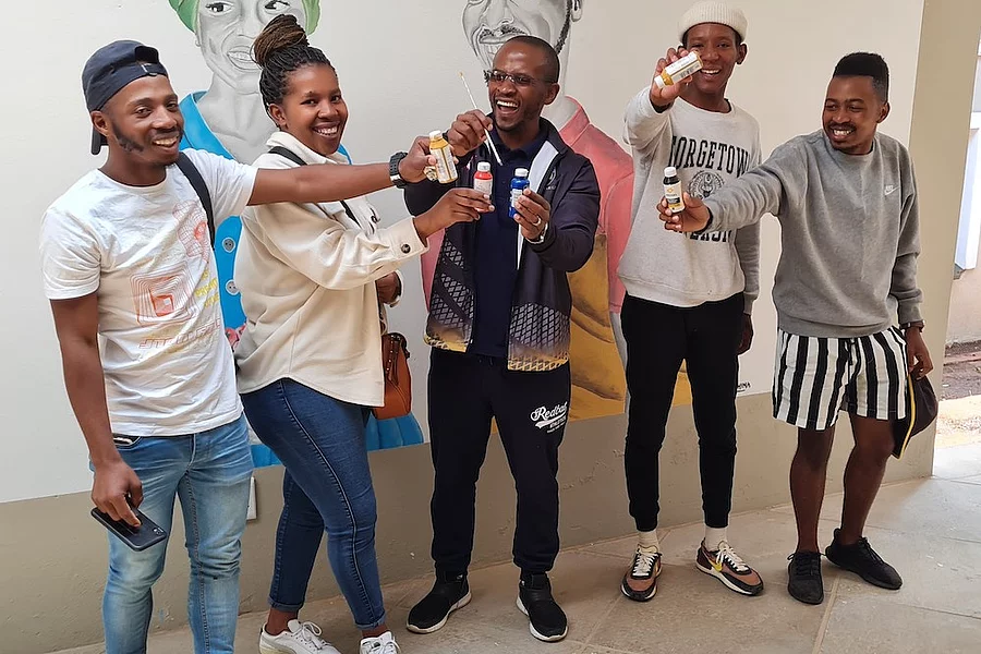 Group of five young people standing indoors in front of a mural featuring handpainted portraits, smiling and holding up small paint bottles.
