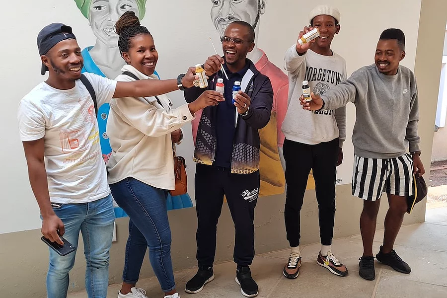 Group of five young people standing indoors in front of a mural featuring handpainted portraits, smiling and holding up small paint bottles.
