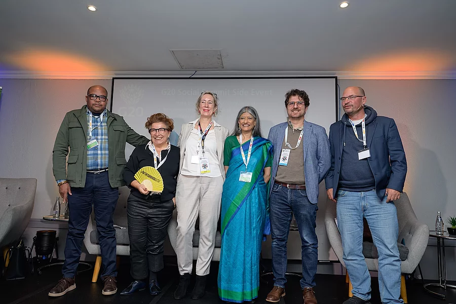 Six speakers (from left:  Tengo Tengela - Trade and Industrial Policy coordinator, COSATU,  Susana Ruiz - Global lead on Tax Justice for Oxfam International, Isobel Frye - Senior Policy Advisor: G20 at Oxfam South Africa, Professor Jayati Ghosh - Co-chair of ICRICT and a member of the G20 Extraordinary Committee of Experts on Global Inequality, Gilad Isaacs - Executive Director, IEJ and Bodo Ellmers - Managing Director Global Policy Forum Europe) side by side on a conference stage, smiling at the camera during a G20 Social Summit side event. Each person wears a conference lanyard, with one holding a yellow hand fan and another dressed in a bright blue and green sari. Behind them is a presentation screen displaying the event title, while armchairs and small tables with water bottles frame the stage, suggesting a panel discussion on public policy or global governance.