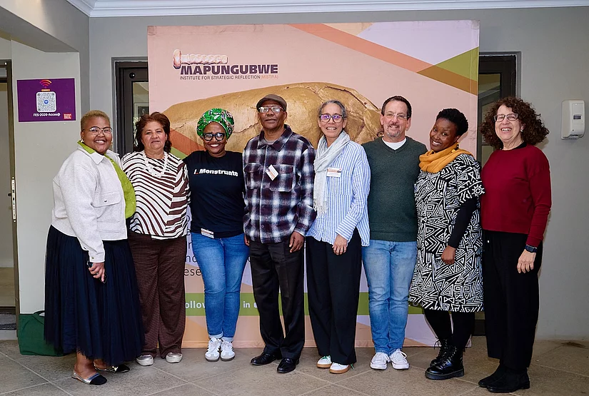 Group photo of eight people standing side by side and smiling, posing indoors in front of a large step-and-repeat banner for the Mapungubwe Institute for Strategic Reflection (MISTRA), featuring an image of the Mapungubwe gold rhino.