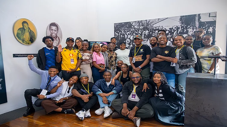 A diverse group of around 20 young people pose together inside a museum or exhibition space with wooden floors and white walls. Some are standing while others sit on the floor, smiling and looking at the camera. Several wear event badges on lanyards. Behind them is a large black-and-white historical photograph and two portrait displays mounted on the wall, creating a sense of learning, heritage, and collective engagement.